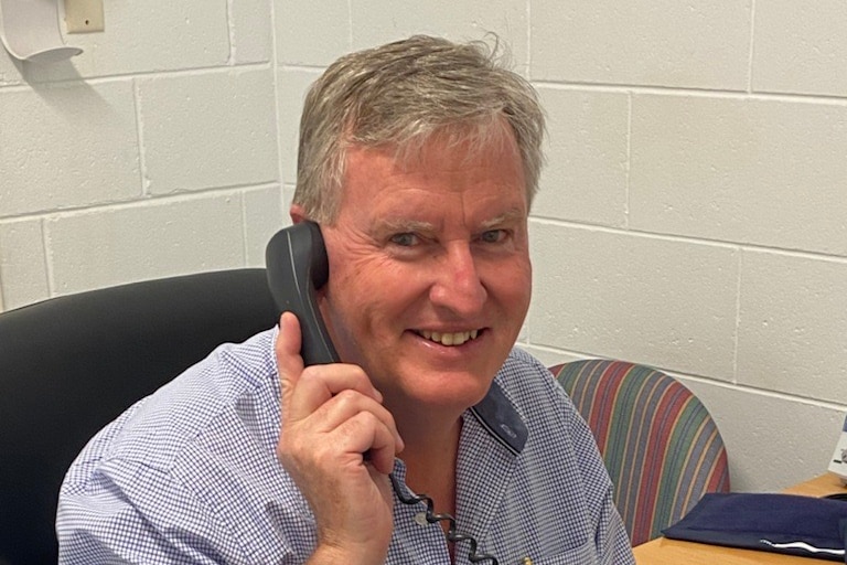 A man with grey hair holds a landline phone receiver to his ear and smiles at the camera.