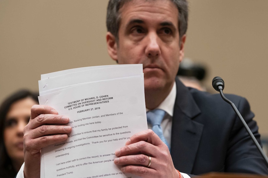 Michael Cohen holds up a sheaf of papers while sitting in front of a microphone.
