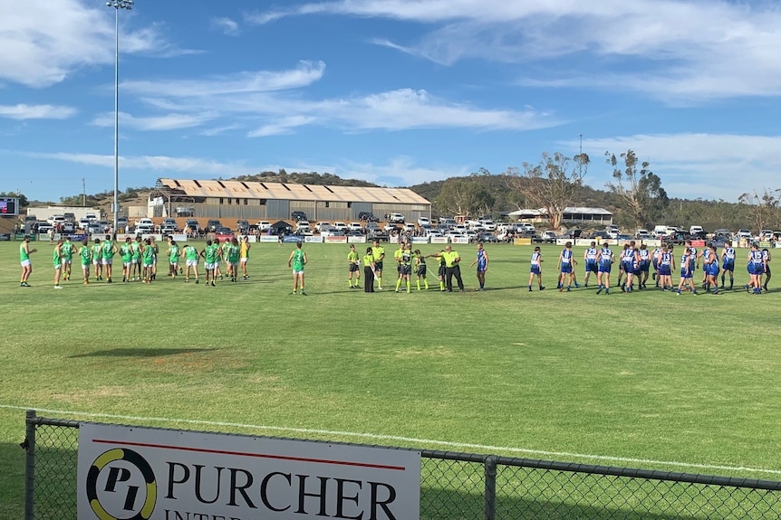 A wide shot of two teams standing on a footy oval on a sunny day. Umpires stand in between them.
