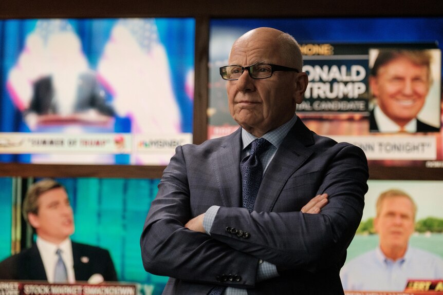 Simon McBurney in character as Rupert Murdoch, a bald middle-aged white man in a dark grey suit standing in a newsroom.