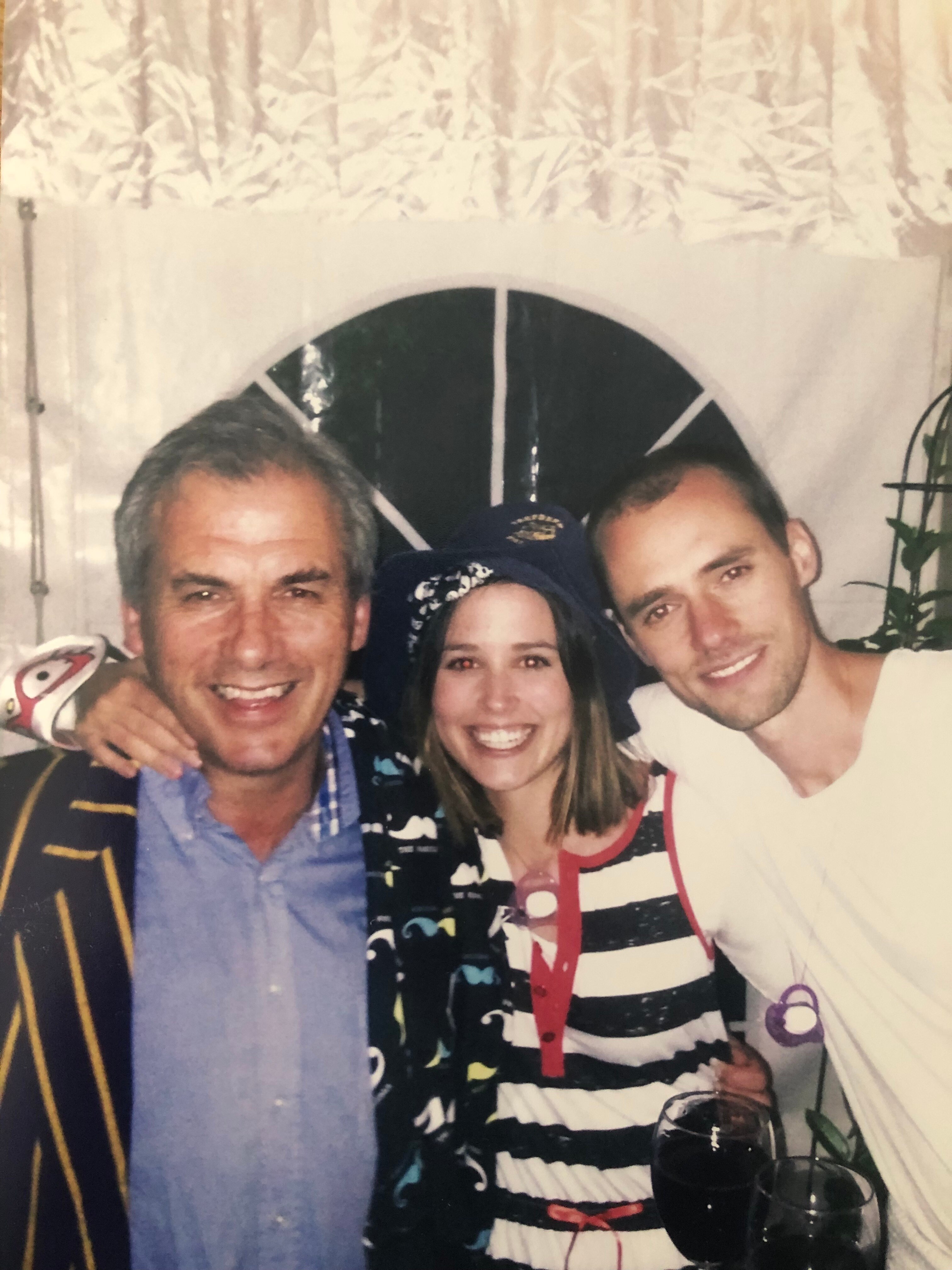 Slightly grainy photo of Rob Moodie, with short grey hair and wide smile, with arms around woman and man, also smiling widely.