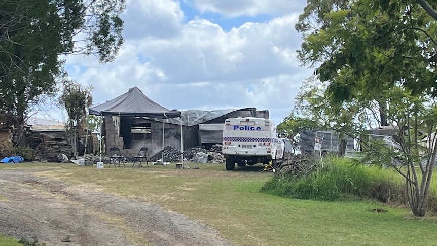 A police van parked at a rural property, a gravel drive way at the front, a burned out structure behind.