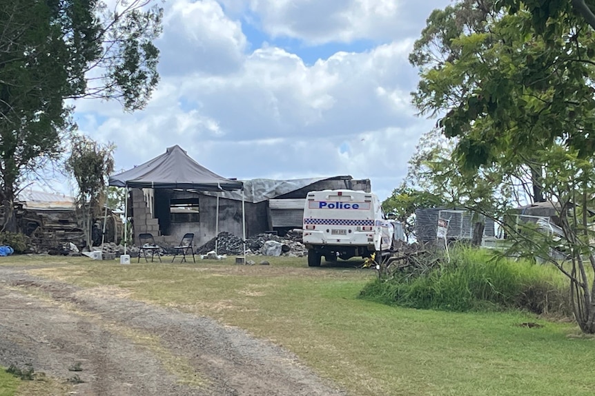 A police van parked at a rural property, a gravel drive way at the front, a burned out structure behind
