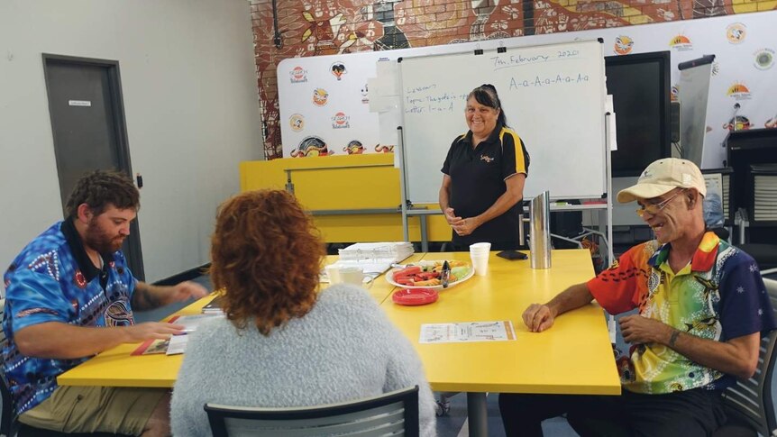 Three people sit around a large yellow table in front of a woman, who stands in front of a whiteboard.