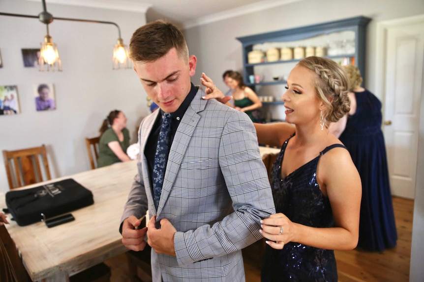 A young man in a grey suit has his collar corrected by a young woman wearing a blue formal dress.