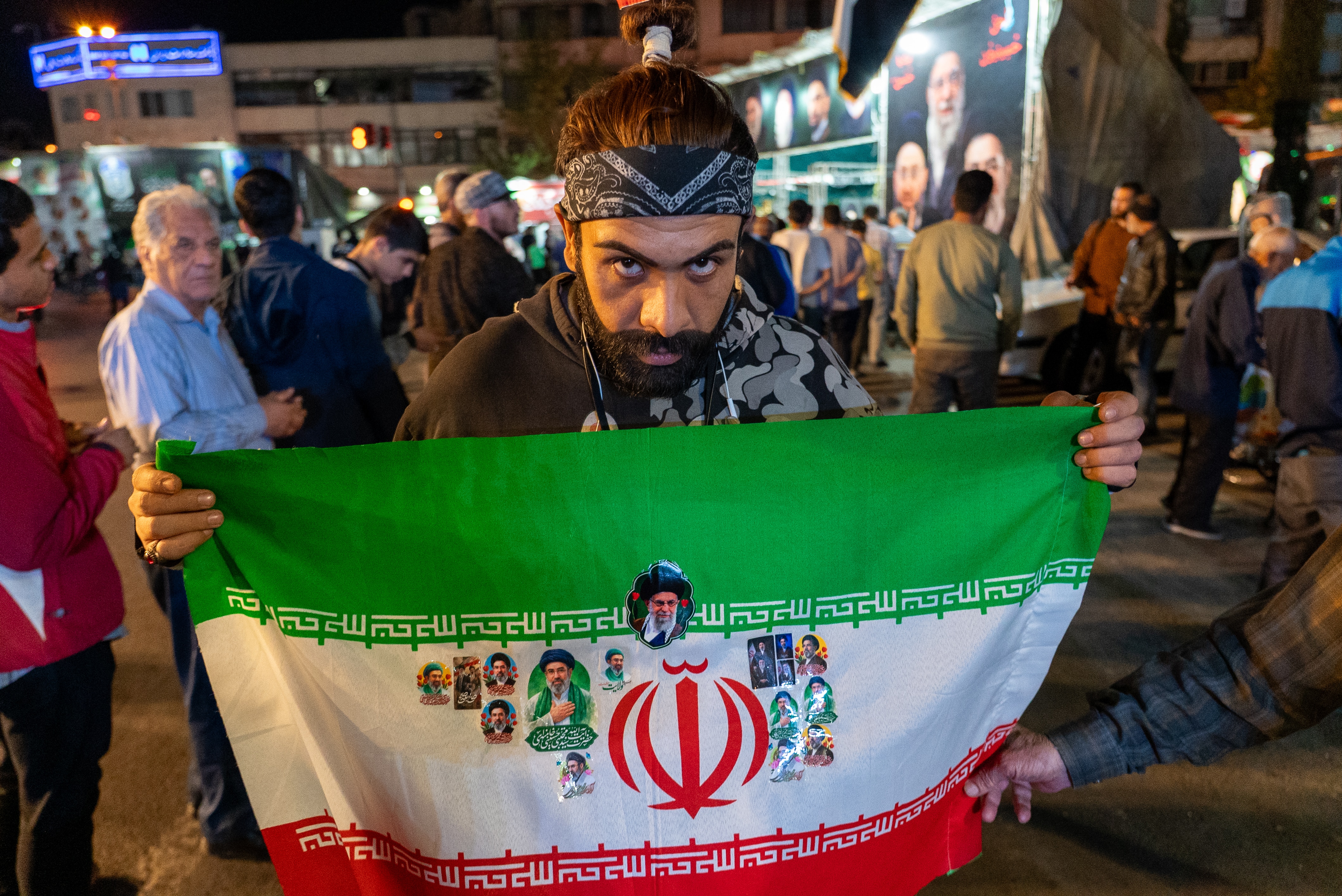 A man wearing a bandana holds up a flag of Iran amongst a crowd.