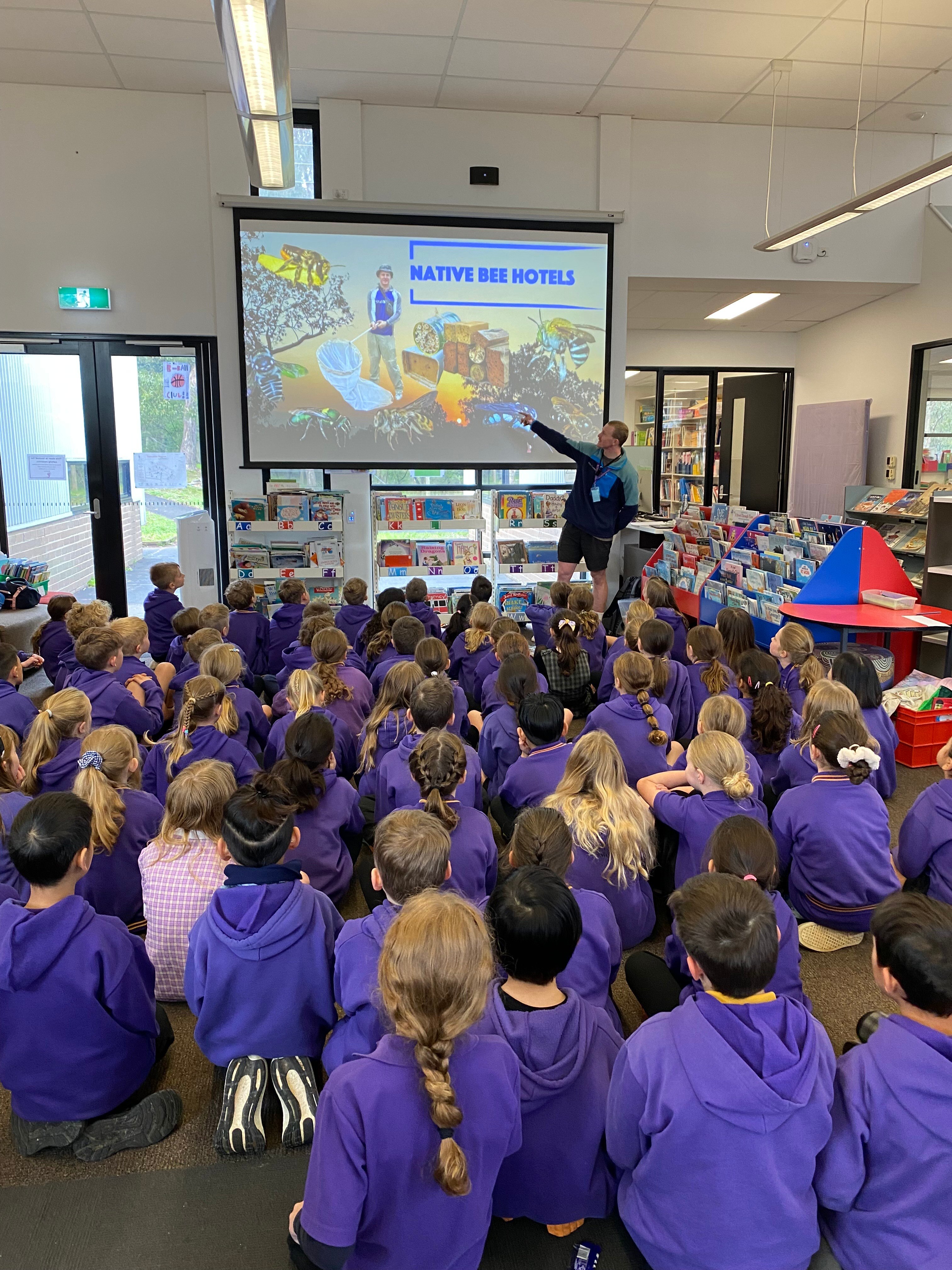 A photo of Clancy pointing at a projector screen, while delivering a workshop on native bee hotels to a room full of students.