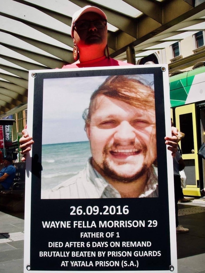 At a bus stop, a woman holds a sign with Wayne's picture stating he died after 6 days on remand.