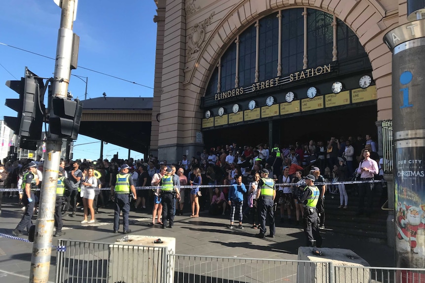 A crowd forms outside Flinders St Station behind police tape. Five police face the crowd.