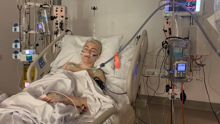 A woman with short bleached hair lays in a hospital bed with her eyes closed, hooked up to a number of tubes and wires.
