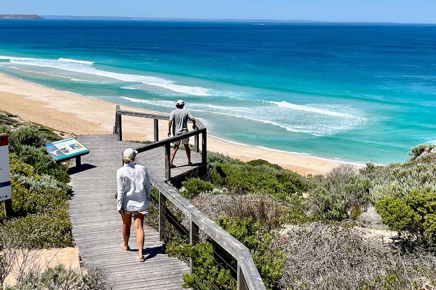 Two people stand on a boardwalk overlooking a long blue beach