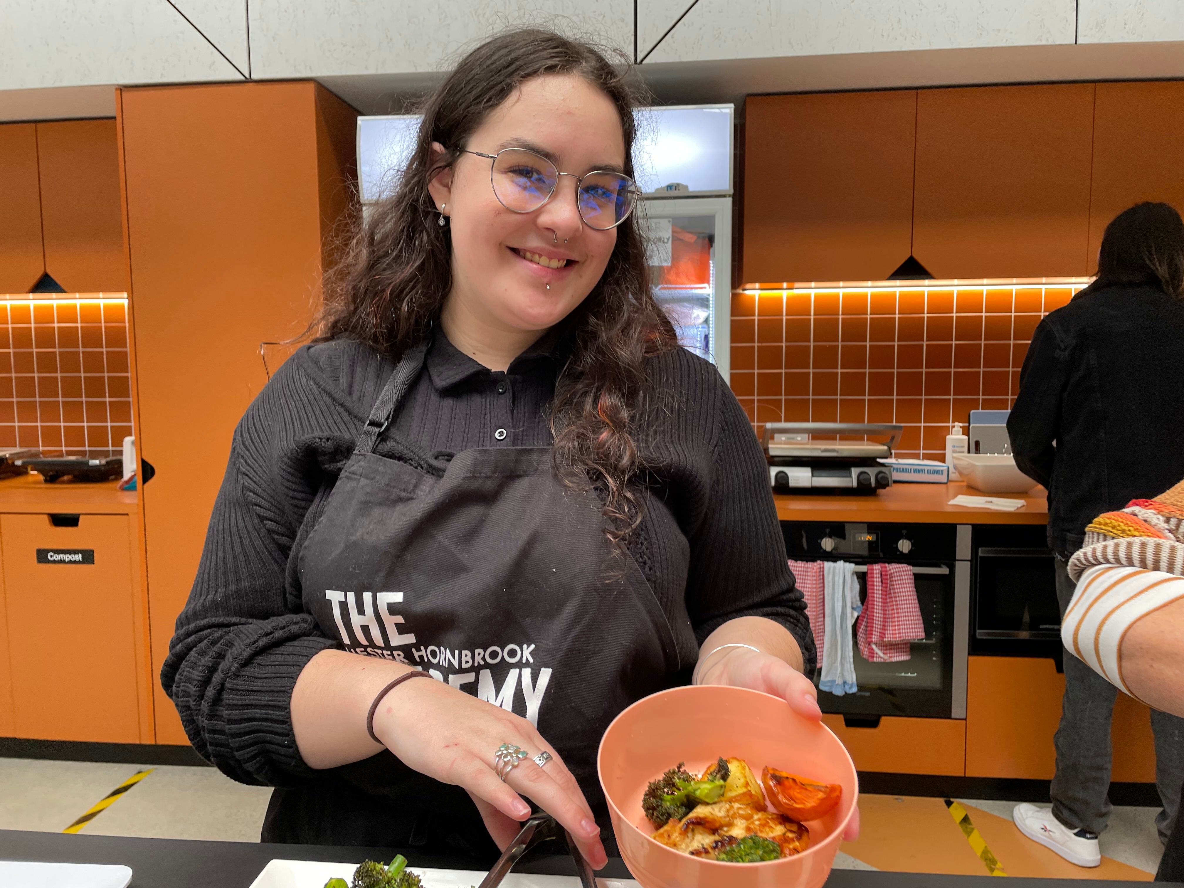 A teenage girl in a black top and glasses smiles as she presents a plate of vegetables while standing in a kitchen. 