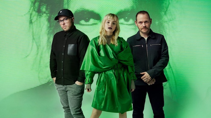 A 2021 press shot of Chvrches against a screen projection of The Cure's Robert Smith