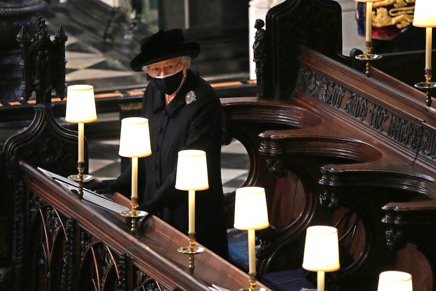 The Queen is dressed in black and sits alone among ornate wooden church pews.