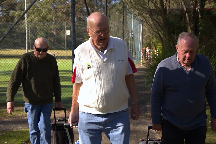 Three men pull small wheeled bags containing their lawn bowls equipment. Behind them is a fenced, grass tennis court.