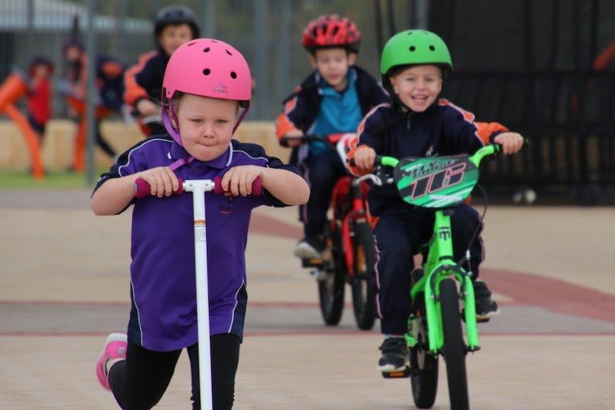 primary school children with helmets on riding scooters and bikes in a school playground
