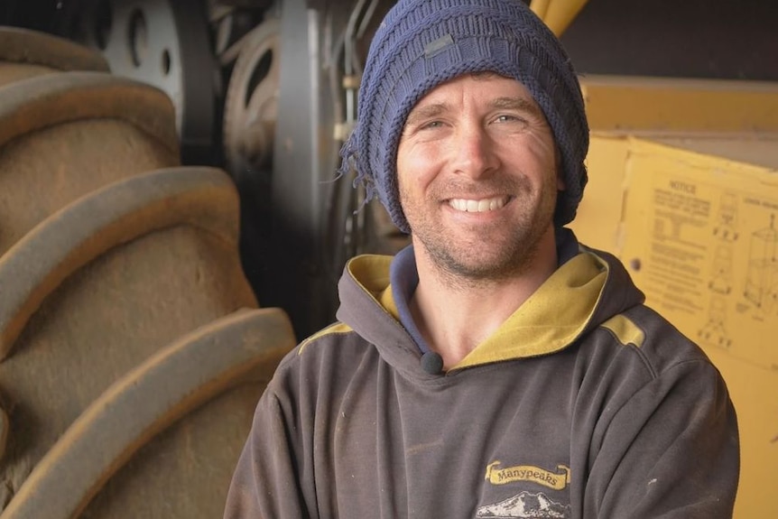 Man in beanie stands in front of big yellow tractor