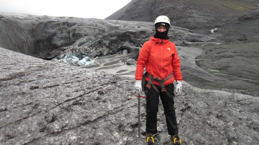 A woman stands covered from head to toe in protective snow gear, wearing spiked shows and a helmet, near glaciers and mountains.