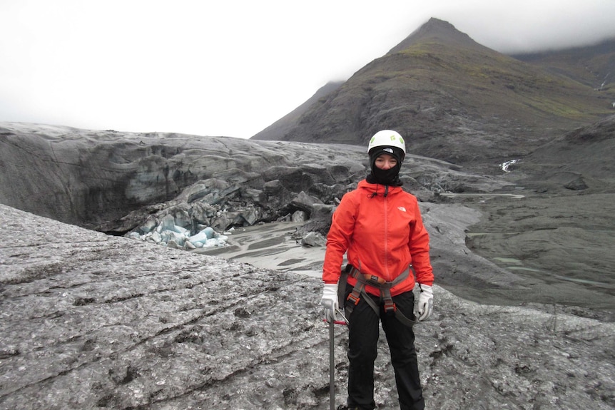 A woman stands covered from head to toe in protective snow gear, wearing spiked shows and a helmet, near glaciers and mountains.