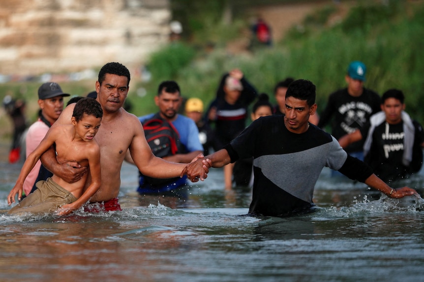 Men, arm in arm, with one carrying a child, walk through a river