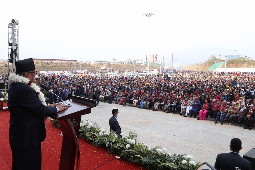 A man stands behind a podium on a stage and speaks to a large crowd. 