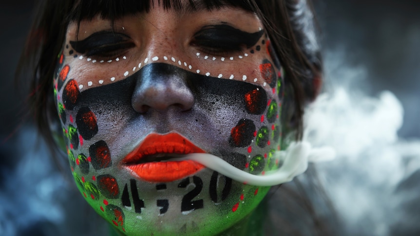 A woman wearing marijuana-themed face paint exhales smoke