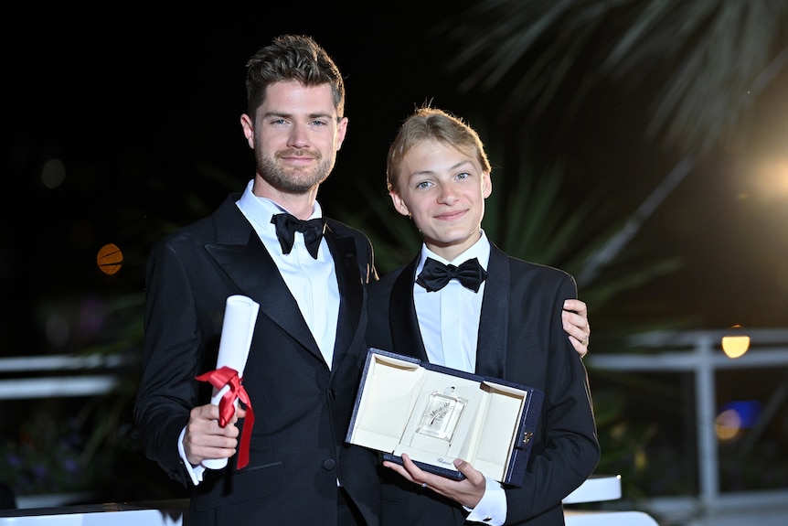 A 30-something white man with short brown hair in a suit stands next to a blond teen boy in a suit, on a red carpet.