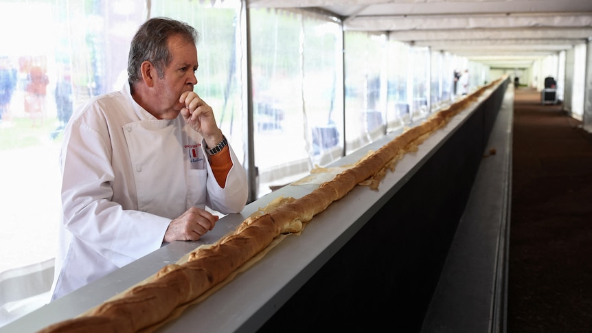 A French baker stands pensively next to a really long baguette coming out of the oven.