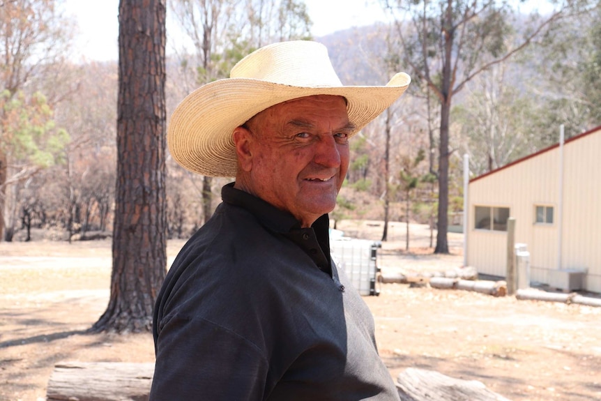 A man in a cowboy hat smiles as he stands on a property with a charred landscape.