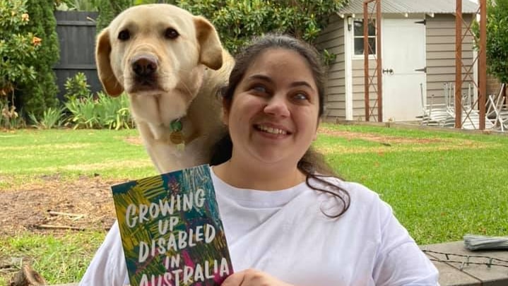 Writer and critic Olivia Muscat stands with her guide dog over her shoulder, holding a copy of "Growing Up Disabled in Australia