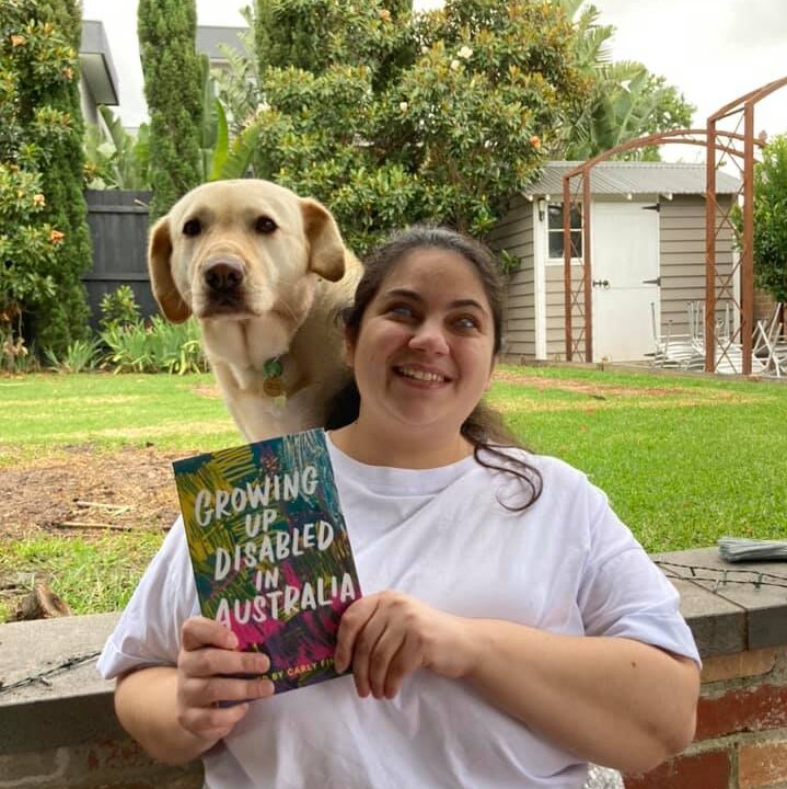 Writer and critic Olivia Muscat stands with her guide dog over her shoulder, holding a copy of "Growing Up Disabled in Australia