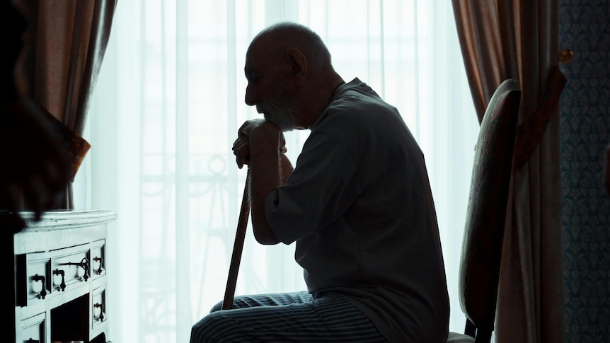 an old man sits on a chair and leans on a walking stick with a desk in front of him and curtains covering a window