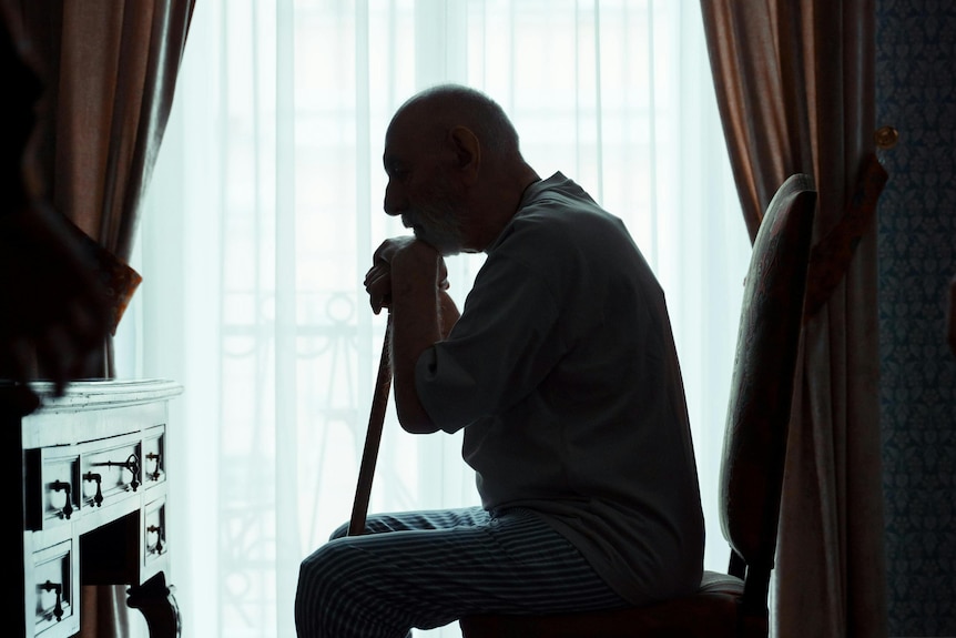 an old man sits on a chair and leans on a walking stick with a desk in front of him and curtains covering a window