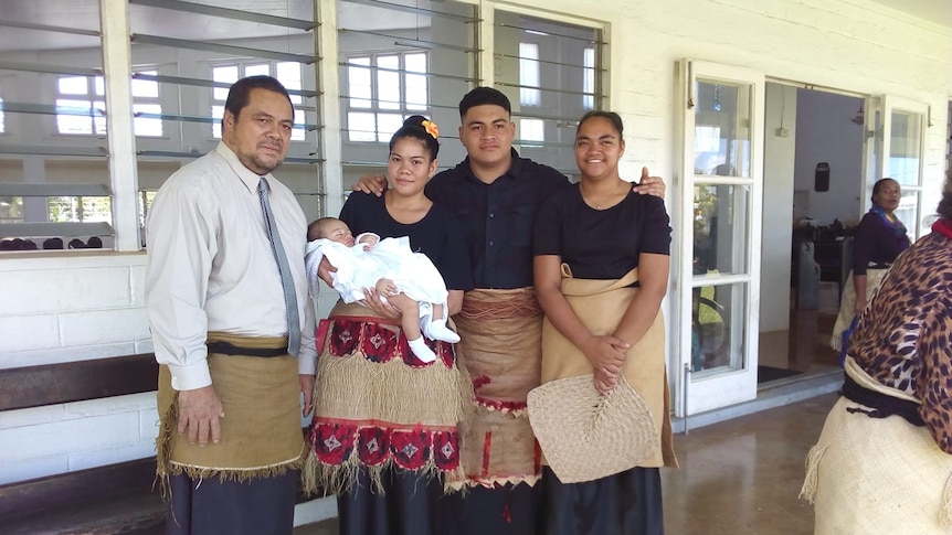 A group of four Polynesian people stand in corridor outside a house with a woman in black holding newborn baby