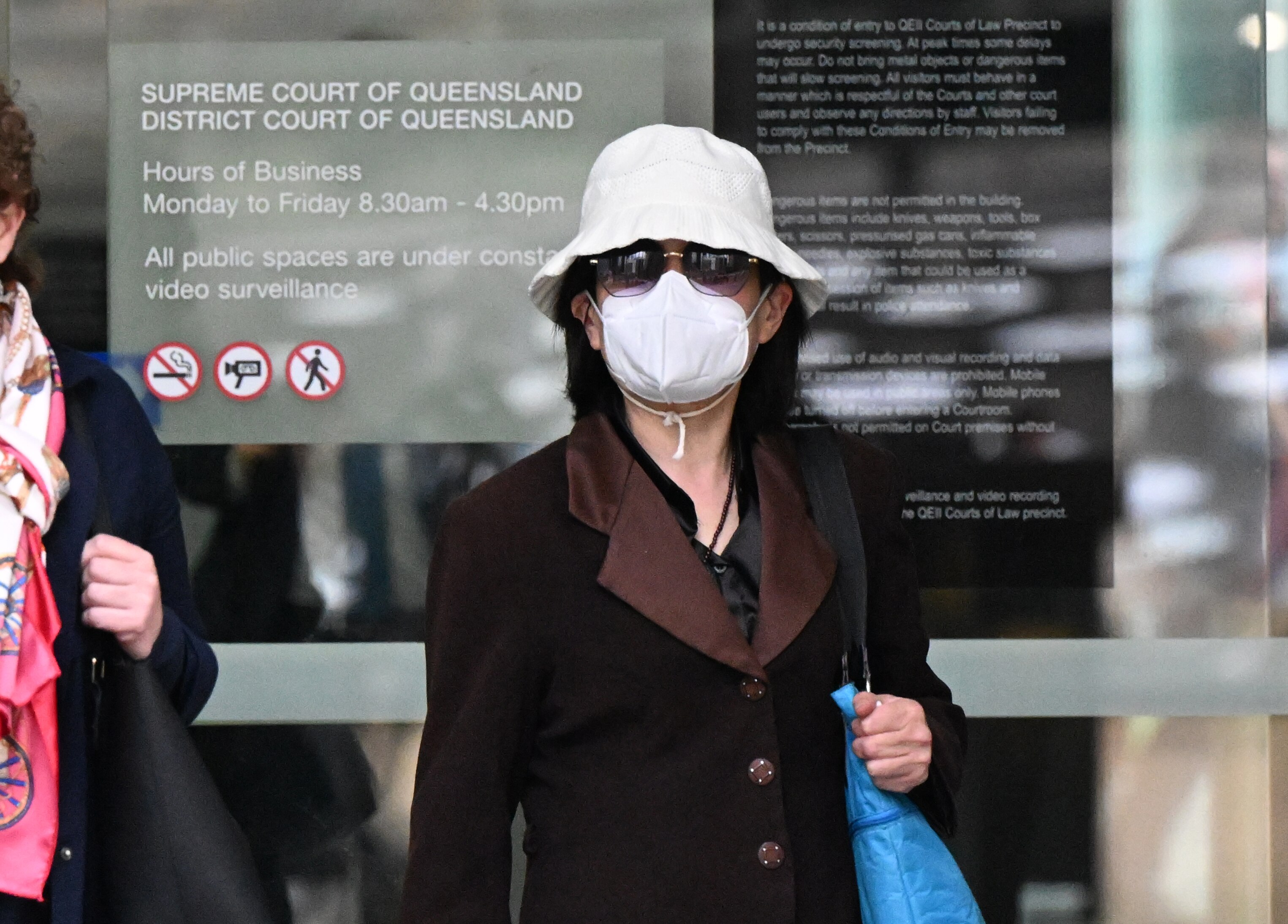 布里斯班最高法院前，一名黑发、身穿深色外套、戴着白色渔夫帽、戴着面具和太阳镜的女子。