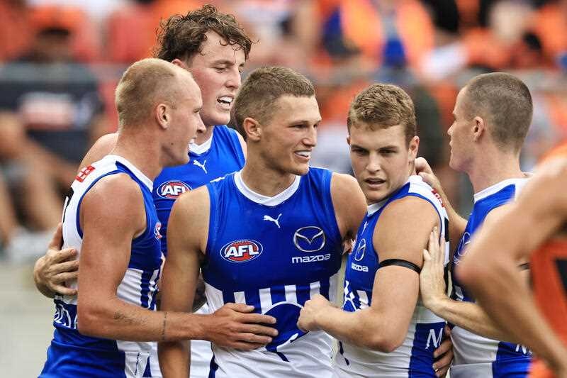 A Kangaroos AFL player smiles as he is surrounded by teammates after kicking a goal. 