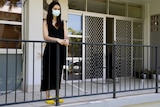 A woman wearing a surgical mask standing on the balcony of an apartment.