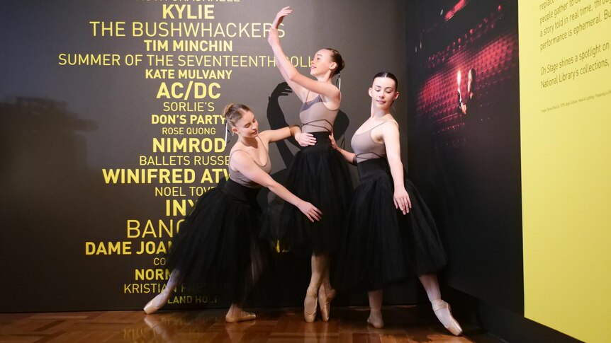 Three young ballerinas stand in front of a wall with the names of famous musicians.