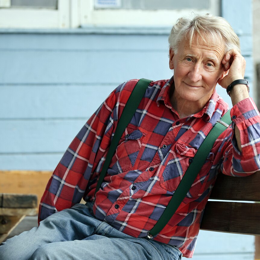 Actor John Gaden wearing a red and blue plaid shirt and green braces and sitting on a bench. 