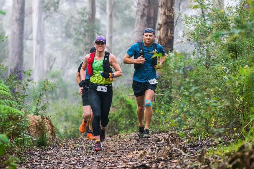 A woman is running through a forest at the head of a pack of runners