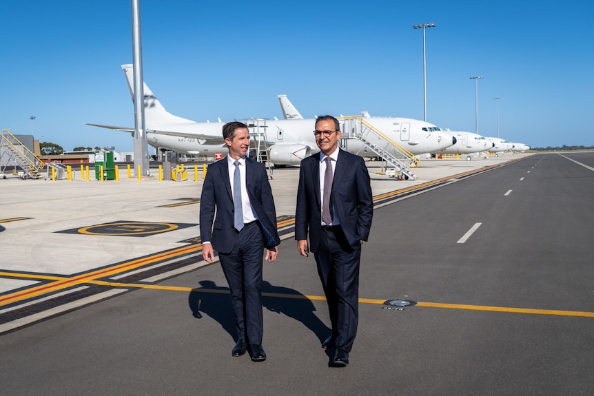 Two men in navy suits walk on a tarmac. Behind them are a series of white planes