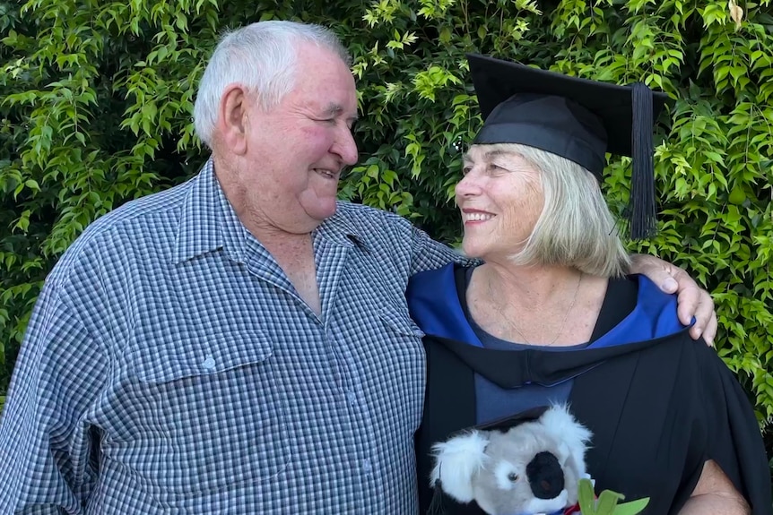 A smiling older woman in a graduation gown and hat hugging her smiling husband in front of green hedge.