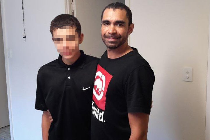 Daniel stands in front of a white wall, his arm around his son, whose face is blurred.