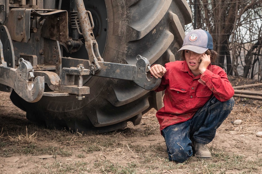 Will Kippel squats down next to the wheel of a tractor, wearing a red shirt, cap and jeans. Dirt covers his face.