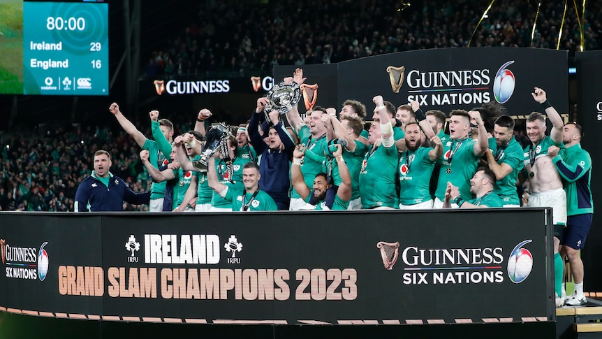 The Irish men's rugby union team celebrate with a trophy behind a sign saying 'Ireland Grand Slam Champions 2023'