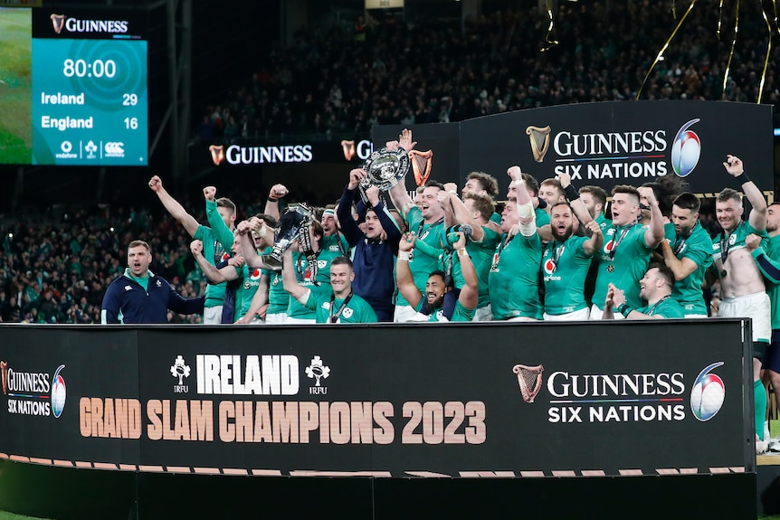 The Irish men's rugby union team celebrate with a trophy behind a sign saying 'Ireland Grand Slam Champions 2023'