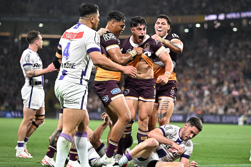 An NRL player is surrounded by teammates after scoring, screaming in excitment to the crowd.