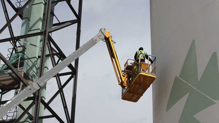 Man in a crane painting big white grain silos with green triangle shapes