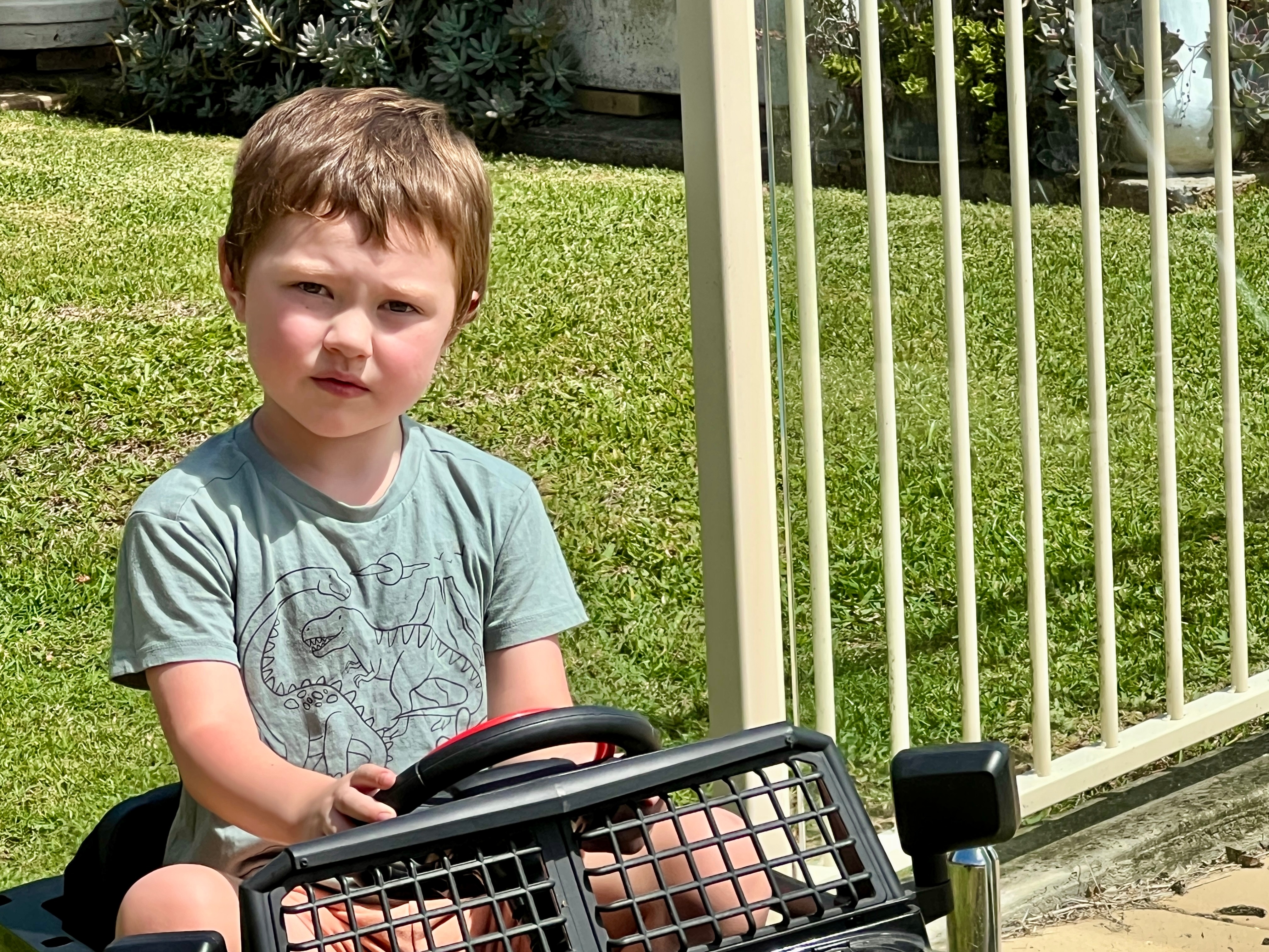 A young boy sits on a toy car in a backyard next to a fence and with green grass behind him.