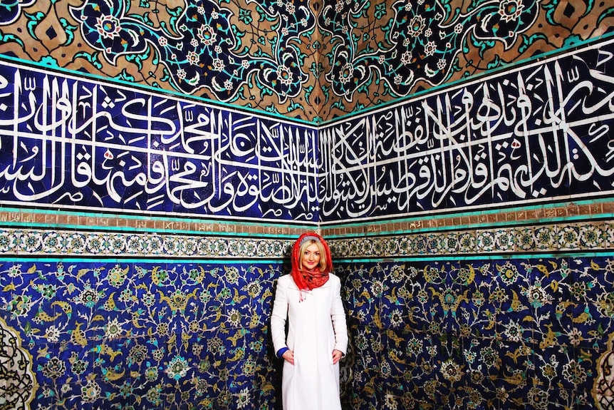 A woman with blonde hair and red head scarf poses in front of a middle eastern building
